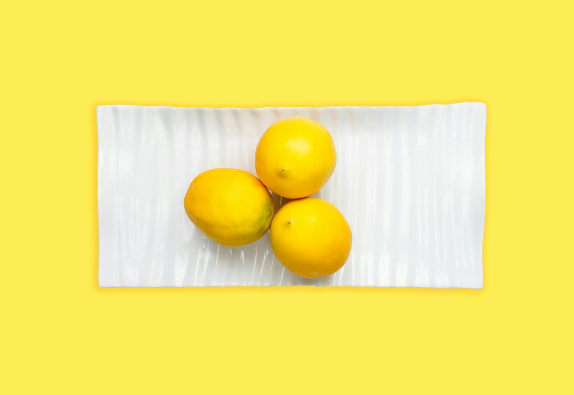 Trois citrons frais arrangés sur une assiette blanche avec un fond jaune vibrant, soulignant la fraîcheur et l'attrait de l'agrume